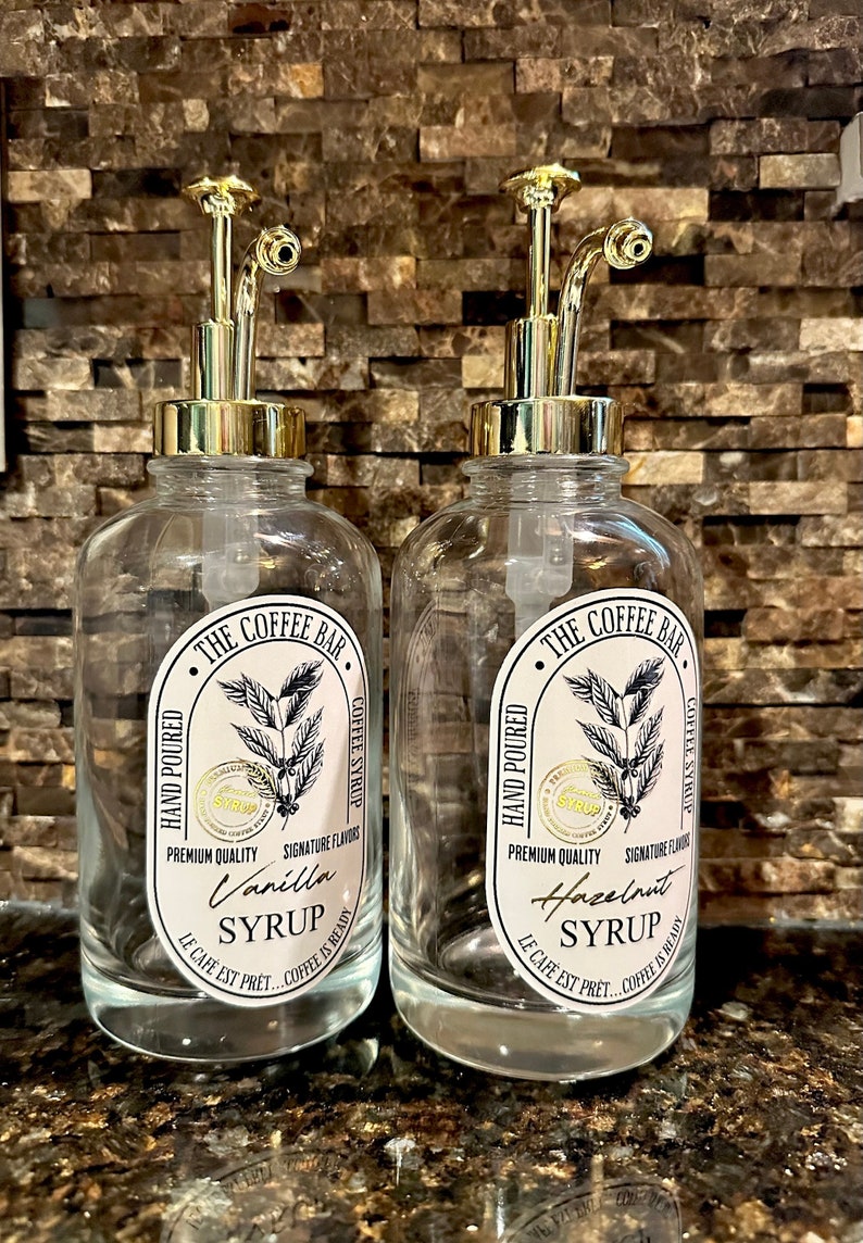 May include: Two clear glass bottles with gold pump tops and labels that say "The Coffee Bar" and feature coffee beans and the text "Hand Poured", "Premium Quality", "Signature Flavors", "Vanilla Syrup", "Hazelnut Syrup", "Le Cafe Est Pret... Coffee Is Ready".