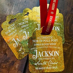 Puede incluir: Etiquetas de regalo doradas con purpurina y cinta roja. Las etiquetas dicen: "Authentic North Pole Post, Express Delivery Service, Special Christmas Delivery to Jackson, Delivered by Santa Claus, The Maker of Holiday Smiles."
