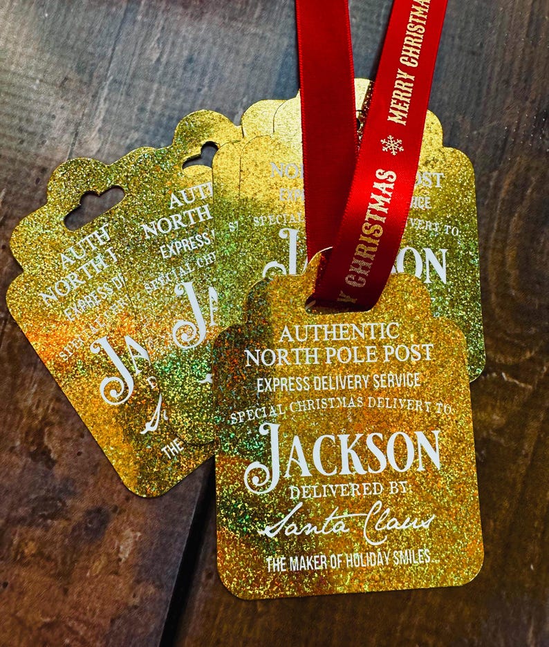 Puede incluir: Etiquetas de regalo doradas con purpurina y cinta roja. Las etiquetas dicen: "Authentic North Pole Post, Express Delivery Service, Special Christmas Delivery to Jackson, Delivered by Santa Claus, The Maker of Holiday Smiles."