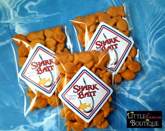 May include: Three clear plastic bags filled with goldfish crackers. Each bag has a diamond-shaped sticker that says "Shark Bait" with a red, white, and blue color scheme and a cartoon fish on a hook.
