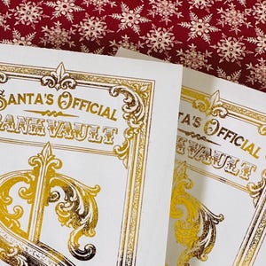 May include: Two white rectangular cards featuring gold foil text that reads Santa's Official Bank Vault. The design includes ornate gold filigree borders and decorative flourishes. The cards are positioned against a dark red background with white snowflakes.