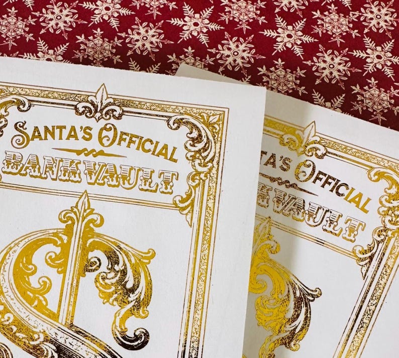 May include: Two white rectangular cards featuring gold foil text that reads Santa's Official Bank Vault. The design includes ornate gold filigree borders and decorative flourishes. The cards are positioned against a dark red background with white snowflakes.