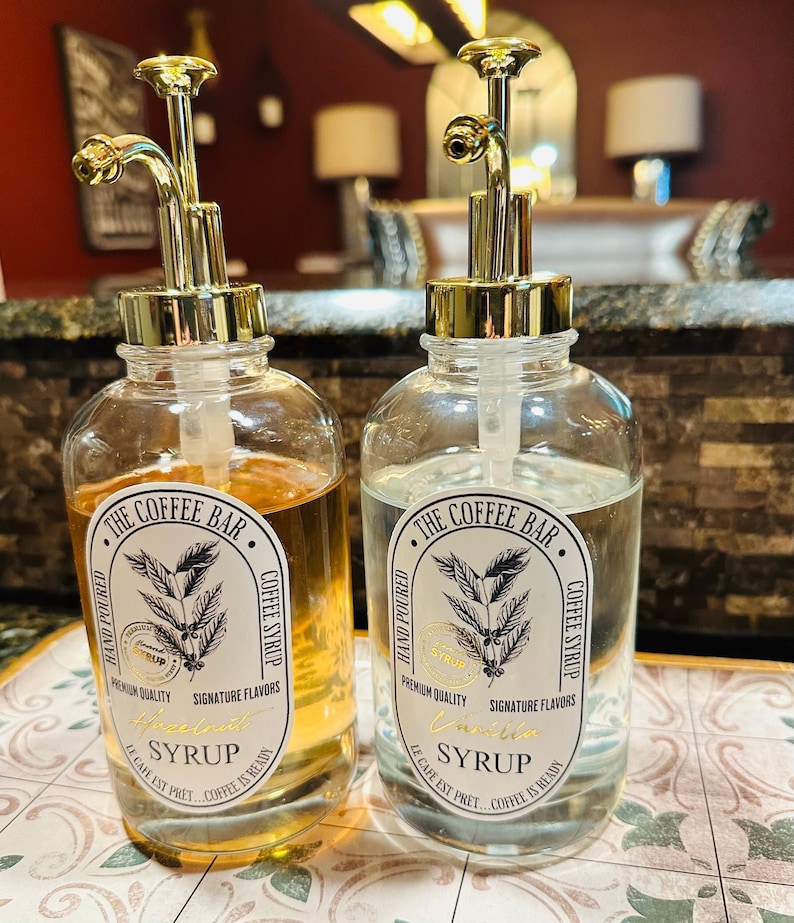 May include: Two clear glass bottles with gold pump tops filled with liquid. The bottles have labels that say "The Coffee Bar" and feature a coffee bean design. One bottle is labelled "Hazelnut Syrup" and the other is labelled "Vanilla Syrup".