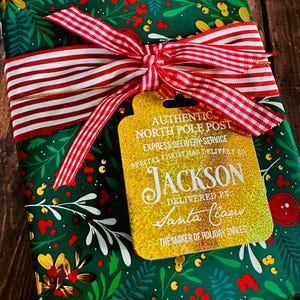 Puede incluir: Un regalo de Navidad envuelto en papel verde con un dise&ntilde;o floral, atado con una cinta a rayas rojas y blancas. Una etiqueta dorada con purpurina dice "AUTHENTIC NORTH POLE POST" y "JACKSON" con "DELIVERED BY Santa Claus."