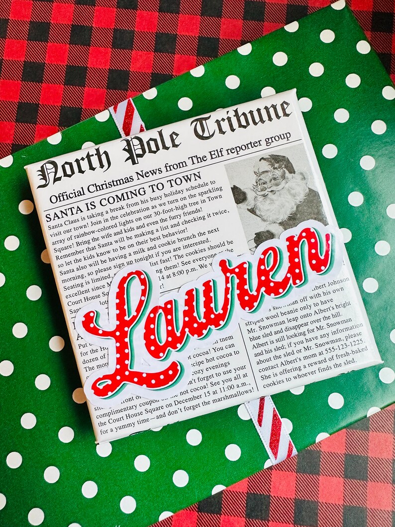 May include: A green and white polka dot wrapping paper with a red and white ribbon. The paper has a newspaper clipping with the headline "North Pole Tribune" and the text "Santa is Coming to Town". There is a red and white sticker with the name "Lauren" on the paper.