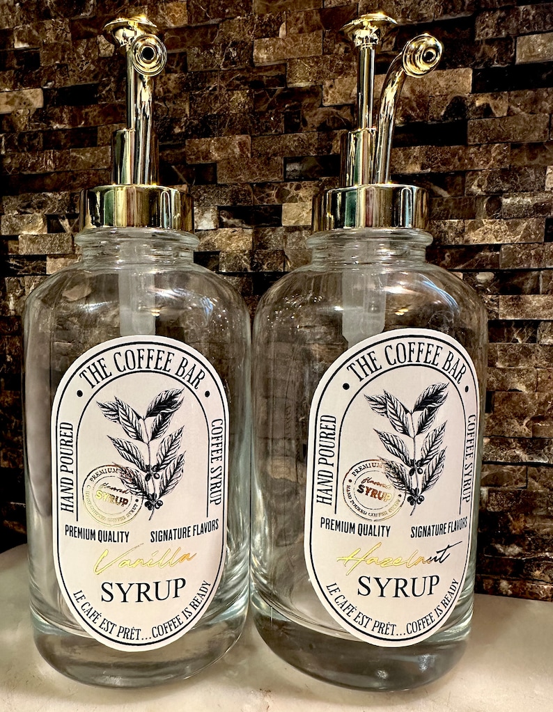 May include: Two clear glass bottles with gold pump tops and labels that say "The Coffee Bar" and feature coffee beans and the text "Hand Poured, Premium Quality, Signature Flavors, Coffee Syrup". One bottle has a label that says "Vanilla Syrup" and the other says "Hazelnut Syrup".