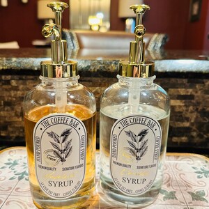 May include: Two clear glass bottles with gold pump tops and labels that say "The Coffee Bar" and "Hand Poured Coffee Syrup". One bottle has a label that says "Hazelnut Syrup" and the other says "Vanilla Syrup".