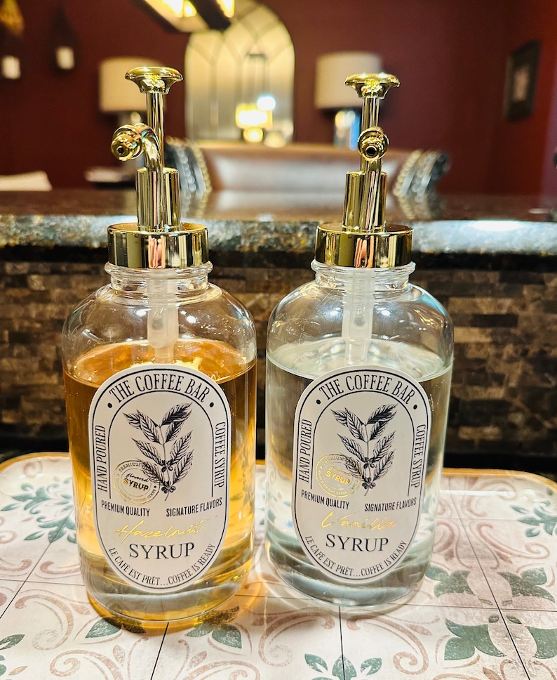 May include: Two clear glass bottles with gold pump tops and labels that say "The Coffee Bar" and "Hand Poured Coffee Syrup". One bottle has a label that says "Hazelnut Syrup" and the other says "Vanilla Syrup".