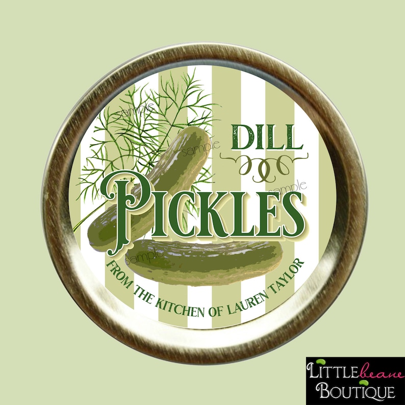 Pu&ograve; includere: Un coperchio rotondo per barattolo dorato con sfondo a strisce bianche e verdi. Il coperchio ha un'etichetta verde e bianca che recita "DILL PICKLES" con un disegno di sottaceti e rametti di aneto. L'etichetta riporta anche "From the Kitchen of Lauren Taylor".