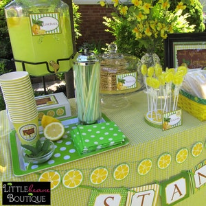May include: A lemonade stand party table setting with a green and white checkered tablecloth. There are green and white polka dot napkins, a glass jar filled with green and white straws, a glass jar filled with lemon-shaped cookies, a glass jar filled with lemonade, and a glass jar filled with lemon lollipops. There is a sign that says "Lemonade Stand" and a banner that says "Stand".