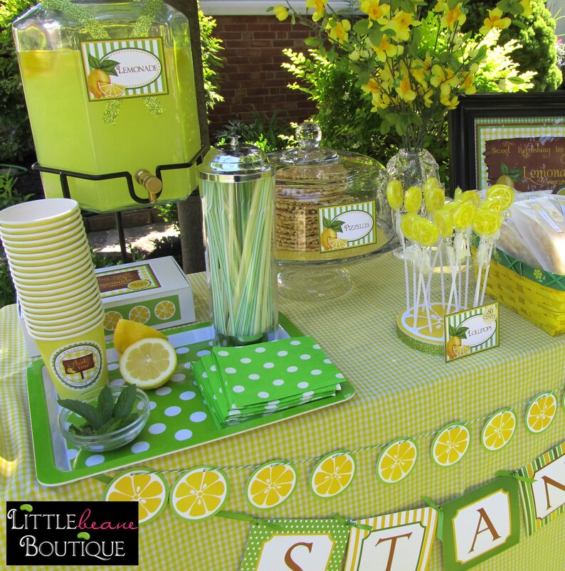 May include: A lemonade stand party table setting with a green and white checkered tablecloth. There are green and white polka dot napkins, a glass jar filled with green and white straws, a glass jar filled with lemon-shaped cookies, a glass jar filled with lemonade, and a glass jar filled with lemon lollipops. There is a sign that says "Lemonade Stand" and a banner that says "Stand".