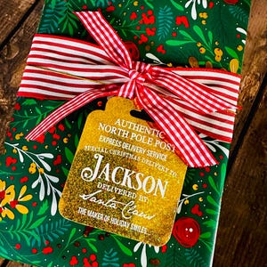 Puede incluir: Un regalo envuelto en papel verde con un dise&ntilde;o floral, atado con una cinta a rayas rojas y blancas. Una etiqueta dorada dice "AUTHENTIC NORTH POLE POST" y "JACKSON DELIVERED BY Santa Claus".