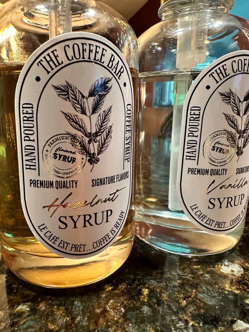 May include: Two glass bottles with pumps, filled with clear liquid. The bottles have labels with gold lettering that reads "The Coffee Bar", "Hand Poured", "Premium Quality", "Signature Flavors", "Hazelnut Syrup", "Le Cafe Est Pret... Coffee Is Ready", and "Vanilla Syrup", "Le Cafe Est Pret... Coffee Is Ready".
