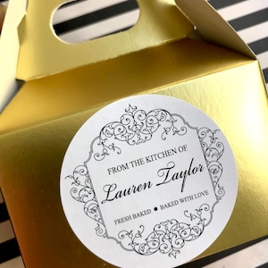 May include: A gold-colored gift box with a handle, featuring a round sticker. The sticker has a decorative border and text that reads "FROM THE KITCHEN OF Lauren Taylor FRESH BAKED • BAKED WITH LOVE."