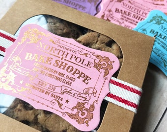 May include: A pink and gold gift box with a window showing chocolate chip cookies. The box has a label that says "Authentic North Pole Bake Shoppe" with the text "Fresh baked by Mrs. Claus and the Christmas Elves" and "Est. 336 Approved by Santa".