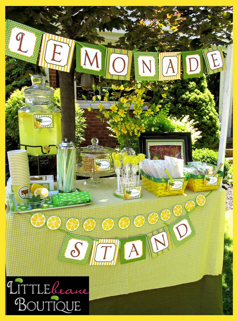 May include: A lemonade stand set up outdoors with a green and yellow checkered tablecloth. The stand is decorated with a banner that reads "Lemonade Stand" and has a pitcher of lemonade, cups, and other treats.