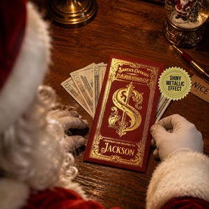 May include: A rectangular red envelope with gold foil detailing, featuring the text Santa's Official Bank Vault and Christmas Cash Delivery To: Jackson, with a large ornate dollar sign in the center, held by gloved hands over US twenty-dollar bills.
