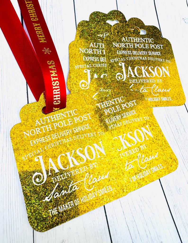 Puede incluir: Etiquetas de regalo doradas y brillantes con el texto "Authentic North Pole Post" y "Jackson" en blanco. Una cinta roja con "Merry Christmas" est&aacute; adjunta. Decoraci&oacute;n navide&ntilde;a.