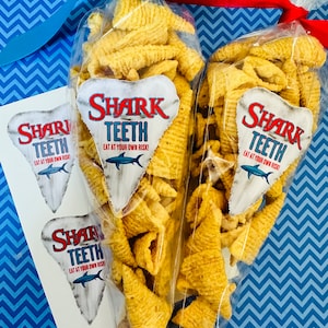 May include: Two clear plastic bags filled with crunchy, golden-brown cone-shaped snacks, tied with a blue ribbon and red ribbon. The bags have white labels with the text "Shark Teeth Eat at your own risk!" printed in red and black.