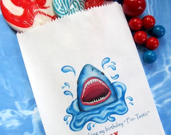 May include: White paper party favor bag with a blue shark illustration and the text "Thanks for making my birthday "Fin-Tastic" ALEX".