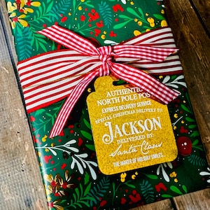 Puede incluir: Un regalo de Navidad envuelto en papel verde con un dise&ntilde;o floral. Una cinta a rayas rojas y blancas est&aacute; atada alrededor del regalo, con una etiqueta dorada que dice "AUTHENTIC NORTH POLE POST EXPRESS DELIVERY SERVICE SPECIAL CHRISTMAS DELIVERY TO JACKSON DELIVERED BY SANTA CLAUS THE MAKER OF HOLIDAY SMILES."