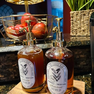May include: Two glass bottles with gold pump tops filled with brown liquid. The bottles have labels with the text "The Coffee Bar" and the words "Vanilla Syrup" and "Hazelnut Syrup".