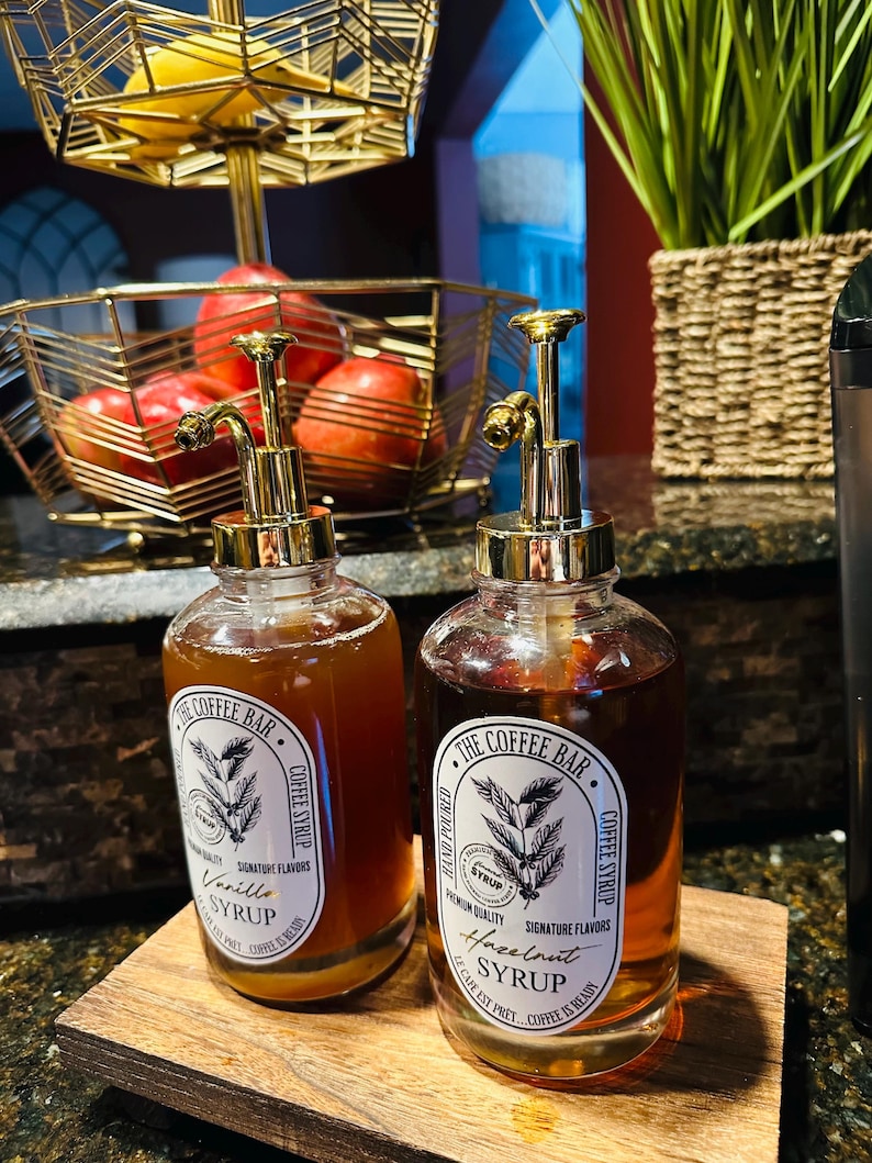 May include: Two glass bottles with gold pump tops filled with brown liquid. The bottles have labels with the text "The Coffee Bar" and the words "Vanilla Syrup" and "Hazelnut Syrup".