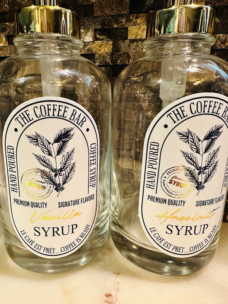 May include: Two clear glass bottles with gold pump tops and labels. The labels are white with black text and a coffee bean illustration. The text on the labels reads "The Coffee Bar", "Hand Poured", "Coffee Syrup", "Premium Quality", "Signature Flavors", "Vanilla Syrup", "Le Cafe Est Pret... Coffee Is Ready" and "Hazelnut Syrup".