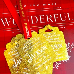 Puede incluir: Etiquetas de regalo doradas con purpurina con el texto "AUTHENTIC NORTH POLE POST" y "JACKSON" en blanco. Una cinta roja con "MERRY CHRISTMAS" est&aacute; drapeada sobre las etiquetas. El fondo es un patr&oacute;n de cuadros rojos y blancos.