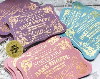 May include: A stack of pink and purple gift tags with gold foil lettering. The tags read "Authentic North Pole Bake Shoppe" with the text "Fresh baked by Mrs. Claus and the Christmas Elves" and "Est. 336 Approved by Santa".
