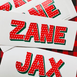 May include: A stack of white stickers with red and white polka dot letters spelling out the names "Zane" and "Jax". The letters have a green outline and a 3D effect.