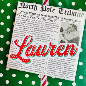 May include: A red and white Christmas-themed gift wrap with a newspaper clipping that reads "North Pole Tribune" and "Santa is Coming to Town". The newspaper clipping also includes a photo of Santa Claus. The gift wrap is decorated with white polka dots on a green background and red and black plaid.