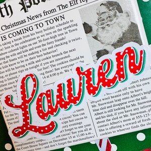 May include: A red and white polka dot sticker with the word "Lauren" in a green and red font. The sticker is on a green and white polka dot background with a red and white plaid background in the corner. The sticker is next to a newspaper clipping with the headline "Christmas News from The Elf reporter group IS COMING TO TOWN".
