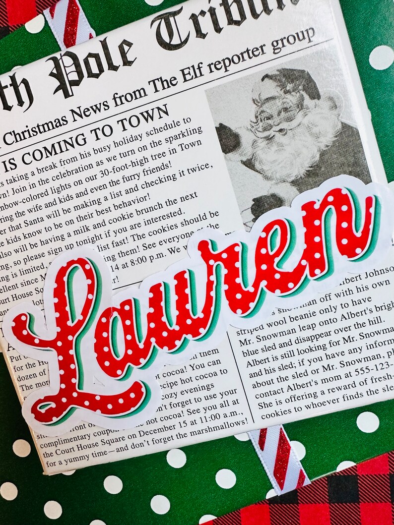 May include: A red and white polka dot sticker with the word "Lauren" in a green and red font. The sticker is on a green and white polka dot background with a red and white plaid background in the corner. The sticker is next to a newspaper clipping with the headline "Christmas News from The Elf reporter group IS COMING TO TOWN".