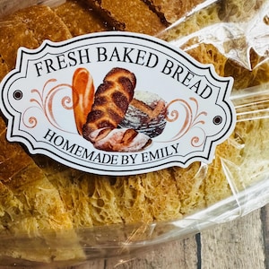 May include: A white label with the text "Fresh Baked Bread Homemade by Emily" and a graphic of three loaves of bread. The label is attached to a plastic bag containing a loaf of sliced bread.