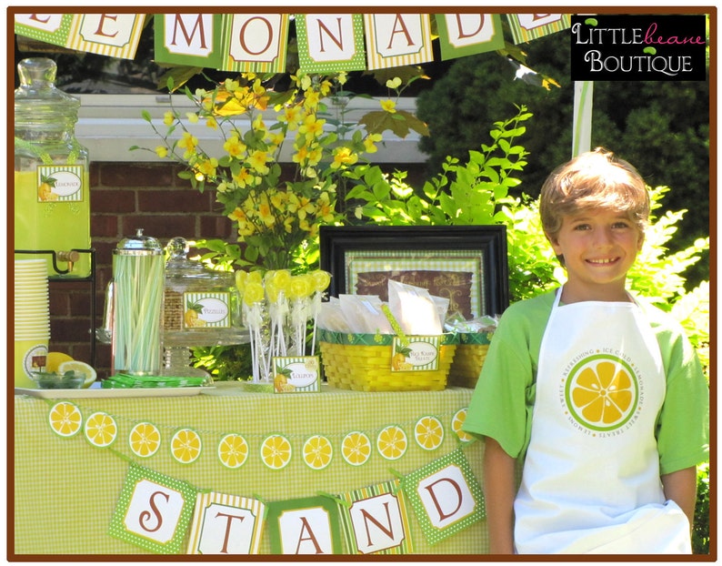 May include: A young boy wearing a white apron with a lemon design and the text "Refreshing & Ice Cold Lemonade" is standing in front of a lemonade stand decorated with yellow and green lemons. The stand has a sign that reads "Lemonade Stand".