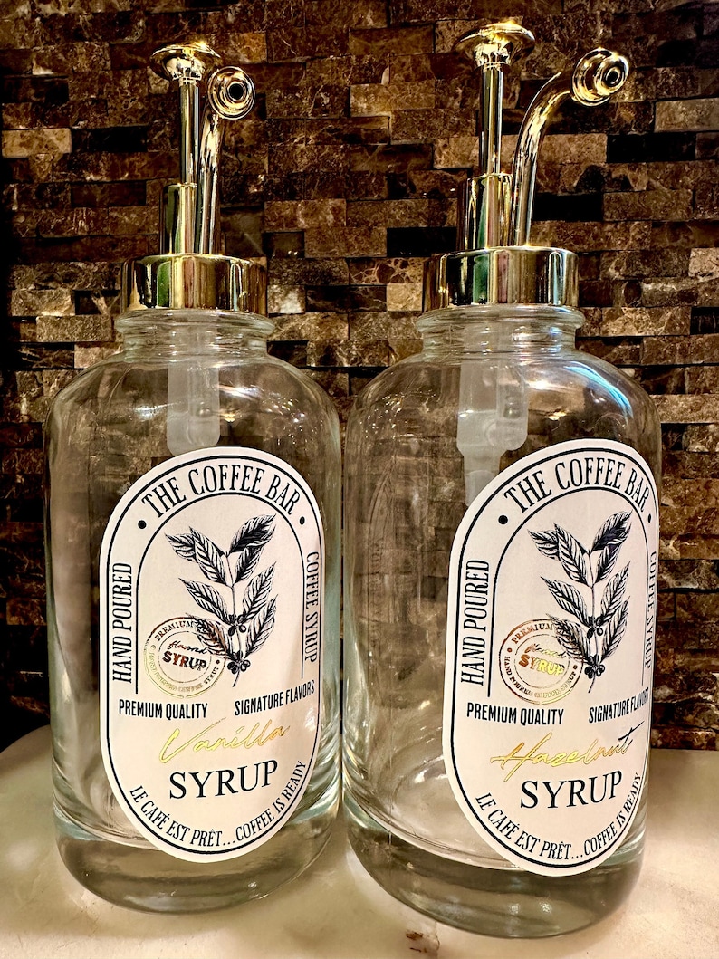 May include: Two clear glass bottles with gold pump tops and labels. The labels are white with black text and a coffee bean illustration. The text on the labels reads "The Coffee Bar", "Hand Poured", "Premium Quality", "Signature Flavors", "Vanilla Syrup", "Hazelnut Syrup", "Le Cafe Est Pret... Coffee Is Ready".