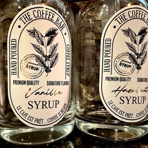 May include: Two clear glass bottles with white labels. The labels feature a black and white illustration of a coffee plant and the text "The Coffee Bar". One bottle says "Vanilla Syrup" and the other says "Hazelnut Syrup".