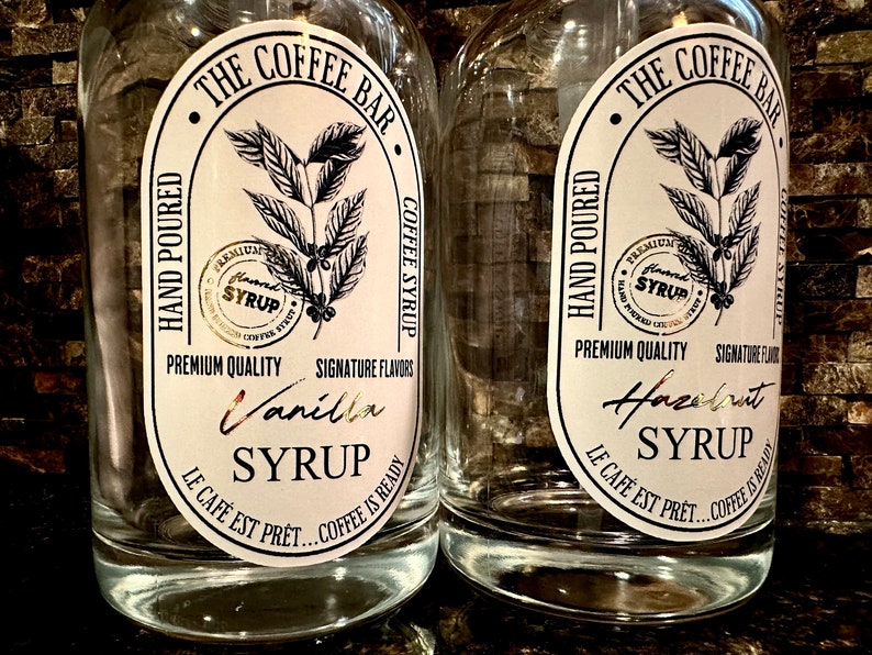 May include: Two clear glass bottles with white labels. The labels feature a black and white illustration of a coffee plant and the text "The Coffee Bar". One bottle says "Vanilla Syrup" and the other says "Hazelnut Syrup".