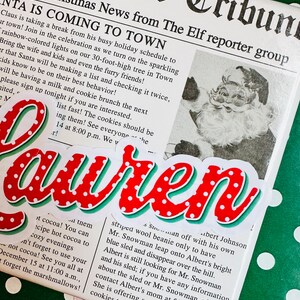 May include: A red and white Christmas-themed newspaper clipping with the headline "Santa is Coming to Town". The newspaper clipping is on a green background with white polka dots. The text on the clipping reads "Official Christmas News from The Elf reporter group".