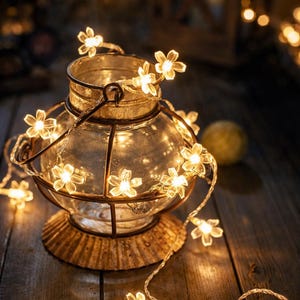 May include: A decorative glass lantern with a metal frame and base, adorned with a string of small, glowing flower-shaped lights. The lantern sits on a wooden surface, creating a warm, inviting ambiance. The lights are a soft, warm white.