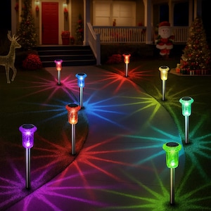 May include: A nighttime scene shows a pathway illuminated by colorful solar garden lights. The lights, with silver poles and various colored tops, cast vibrant patterns on the ground. The scene is set in front of a house decorated for Christmas.