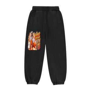 May include: Black sweatpants featuring an elastic waistband with a drawstring and a graphic print on the left leg. The graphic depicts a person in orange clothing with the text Inner Radiance and Owning The Night in yellow and orange lettering.
