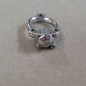 May include: A small, silver-colored ring charm with a large, faceted gemstone. The ring has a classic engagement ring design, with a round band and a prominent central stone. The charm has a small loop at the top for attaching to a necklace or bracelet.