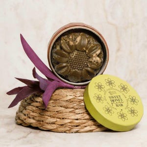 Può includere: Una barra per il trattamento dei capelli di colore marrone scuro a forma di girasole, contenuta in un barattolo circolare. Il coperchio giallo riporta la scritta Sweet Honey Gloss Bar e illustrazioni floreali. Una foglia viola è accanto.