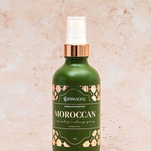 Può includere: Un flacone spray verde bosco opaco con un colletto in oro rosa metallizzato e un tappo trasparente. L'etichetta riporta il testo Henna Sooq, Moroccan Collection, Moroccan infused finishing spray, hydrating anti-frizz e 118 ml in bianco e oro rosa.