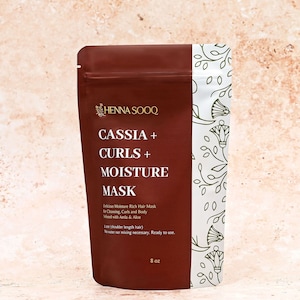 Può includere: Una busta marrone di Henna Sooq Cassia + Curls + Moisture Mask, 8 oz. Il fronte presenta un testo bianco che descrive il prodotto come una maschera per capelli idratante infusa con Amla e Aloe, con un pannello laterale bianco a motivi floreali verdi.