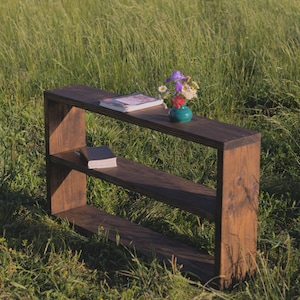 May include: A dark brown wooden bookshelf with two shelves, set outdoors in a field of tall green grass. A book and a small vase of flowers sit on the top shelf, and another book rests on the lower shelf.