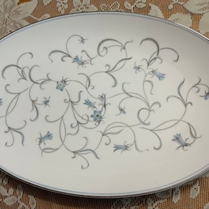 May include: Oval serving platter with a white background and a delicate floral design. The pattern features gray and light blue stylized flowers and vines. The rim is trimmed with a silver band. A classic piece for serving.