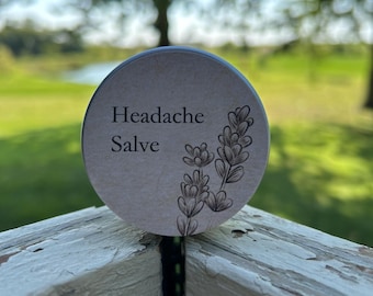 May include: A small, round tin with a brown label that reads "Headache Salve". The label also features a black and white illustration of a lavender flower.